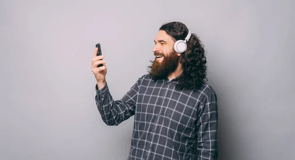 Felice uomo hipster barbuto con i capelli lunghi ascoltando musica a cuffie bianche senza fili e guardando smartphone — Foto Stock