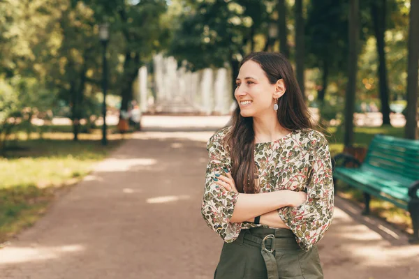 Retrato de jovem feliz com braços cruzados em pé no parque e olhando para longe — Fotografia de Stock