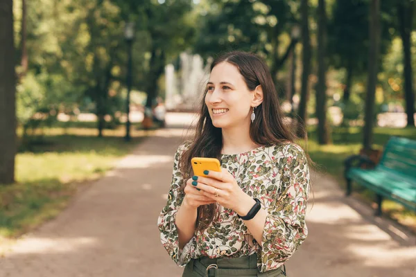 Foto da jovem mulher de pé no parque e segurando smartphone amarelo — Fotografia de Stock