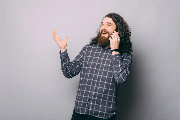 Fiatal szakállas férfi kockás ingben beszél okostelefonon és gesztikulál szürke háttér — Stock Fotó