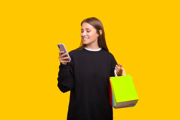 Portret szczęśliwej młodej kobiety trzymającej torby na zakupy i telefon komórkowy na żółtym tle — Zdjęcie stockowe