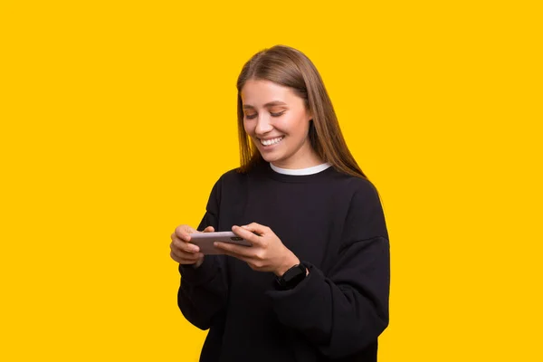 Feliz bonito bela jovem jogar jogos por telefone celular isolado sobre fundo de parede amarela. — Fotografia de Stock