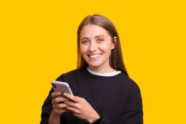 Mutlu güzel bayan cep telefonu kullanıyor, kameraya gülümsüyor, sarı arka planda duruyor..