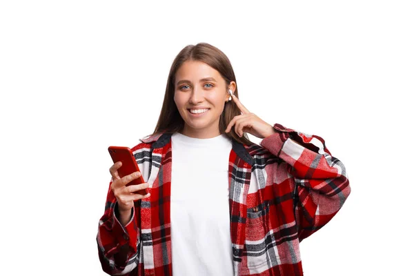 Очаровательная молодая женщина в казуальте, позирует изолированно на белом фоне, слушает музыку с воздушными стручками, держит мобильный телефон — стоковое фото