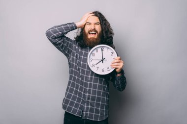 Büyük bir saat tutan yakışıklı sakallı genç adam gergin ve hayal kırıklığına uğramış, şaşırmış ve kızgın yüzlü.