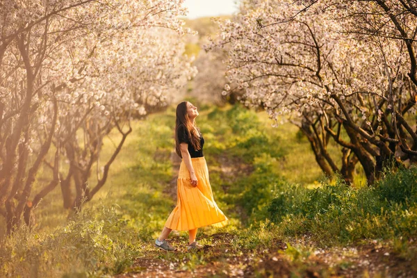 Młoda kobieta ciesząca się wiosną na zielonym polu z kwitnącymi drzewami na zachodzie słońca — Zdjęcie stockowe