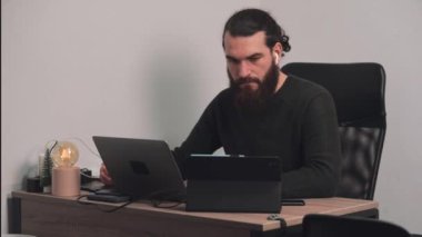 Genç sakallı adamın ofis masasında oturup dizüstü bilgisayarın başında çalışırken çekilmiş görüntüleri.