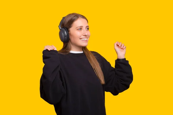 Hermosa chica, escuchando música en auriculares inalámbricos, sonriendo y bailando, de pie sobre fondo amarillo —  Fotos de Stock