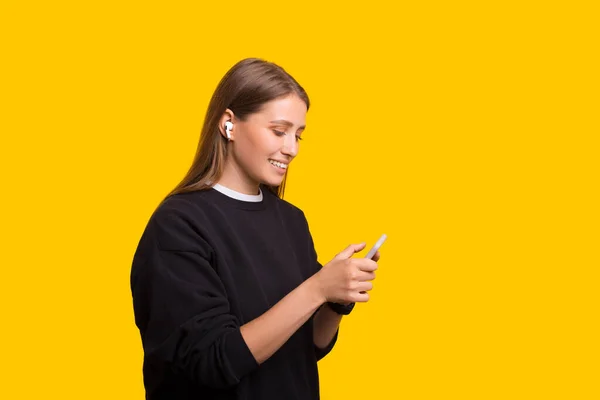 Fiatal nő kezében mobiltelefon hallgat zenét fülhallgató elszigetelt sárga háttér stúdió — Stock Fotó