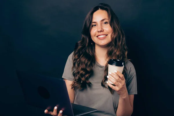 Портрет улыбающейся бизнесвумен с ноутбуком и чашкой кофе, чтобы пойти, на темном фоне — стоковое фото