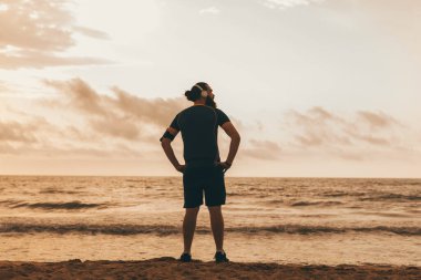 Sahilde durup gün batımını izleyen genç bir adamın arka görüntüsü.
