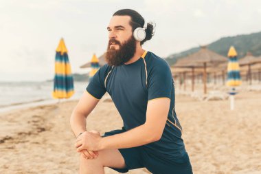 Genç sakallı adam sabah koşusundan önce ısınırken kulaklıkla müzik dinliyor.