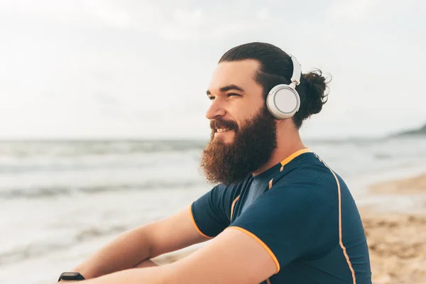 Jeune homme sportif barbu de remise en forme écoutant de la musique au repos regardant le lever du soleil sur la mer — Photo