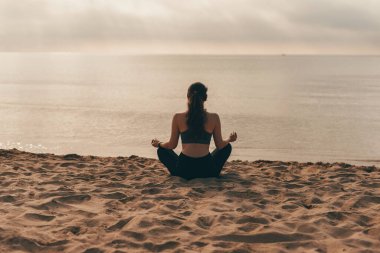 Sabahları sahilde yogacı bir kadın, Yoga Lotus meditasyon pozisyonu almış bir kız gün doğumunda sahilden sahile doğru.