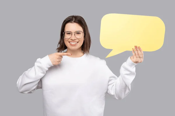 Charming young woman wearing white sweater and pointing at blank yellow speech bubble — Fotografia de Stock