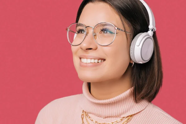 Close up portrait of young beautiful woman smiling and listening music at white wireless headphones over pink background — 스톡 사진