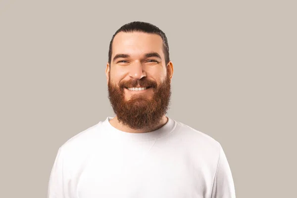 Közelkép portré jóképű fiatal szakállas hipszter férfi néz a kamerába, és mosolyog át szürke háttér — Stock Fotó