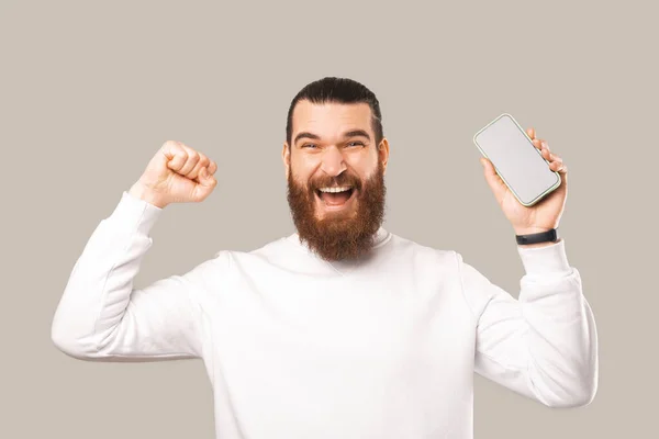 Incroyable jeune homme barbu en sweat à capuche blanc célébrant le succès et tenant smartphone avec écran blanc — Photo