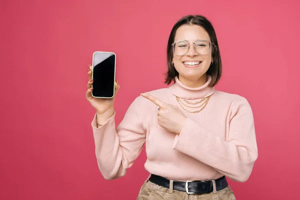 쾌활하고 쾌활 한 젊은 여성 이 핑크 색 배경 위로 스마트폰 화면을 가리키며 웃고 있습니다 — 스톡 사진