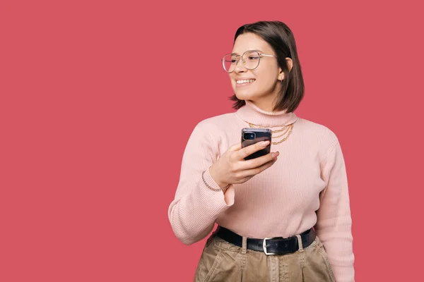 Vidám fiatal gyönyörű nő használja okostelefon felett rózsaszín háttér és nézi copyspace — Stock Fotó