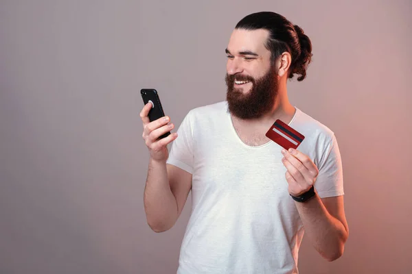 Feliz joven guapo barbudo hombre compra en línea con tarjeta de débito y teléfono inteligente — Foto de Stock