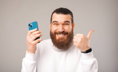 Yakışıklı, sakallı bir adamın baş parmağını kaldıran ve elinde telefon tutan portresini yakından çek..