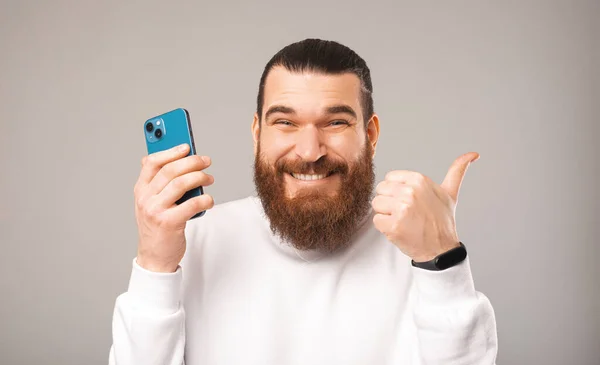 De cerca retrato de un hombre barbudo guapo mostrando el pulgar hacia arriba y sosteniendo un teléfono. — Foto de Stock