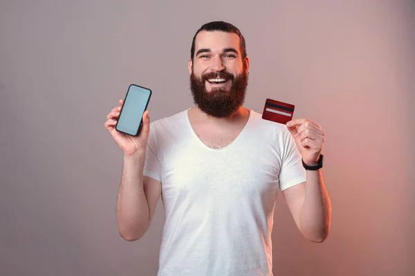 Hombre barbudo alegre está sosteniendo su teléfono y tarjeta de crédito. — Foto de Stock