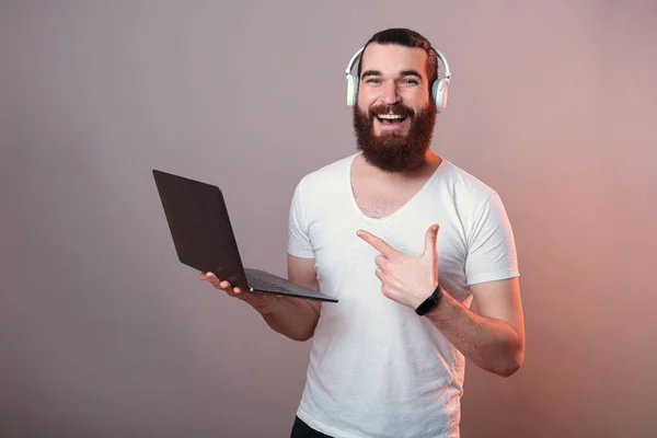 Amplio hombre sonriente está apuntando a la computadora portátil mientras usa auriculares. —  Fotos de Stock