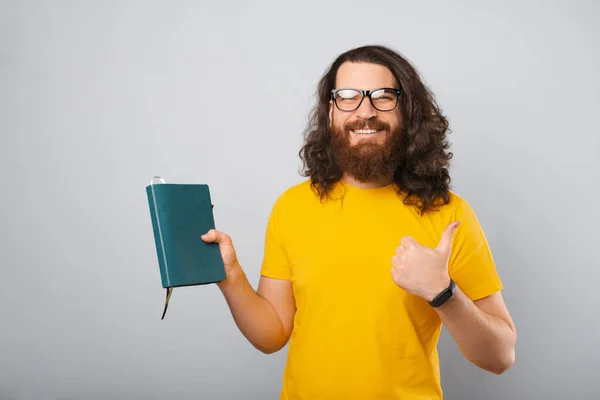 Chytrý mladý muž ukazuje palec nahoru, zatímco drží svůj deník. — Stock fotografie