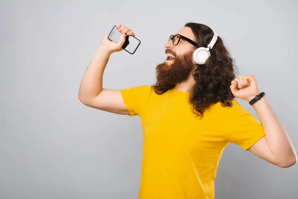 Hipster de pelo largo con auriculares está cantando mientras sostiene el teléfono inteligente. —  Fotos de Stock