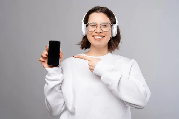 Cute young woman wearing headphones is pointing at the phone she is holding. — Stock Photo, Image