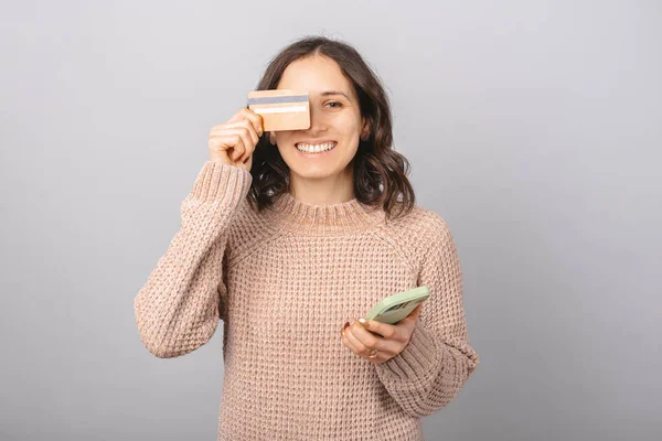 Podekscytowana młoda dama zakrywa jedno oko kartą kredytową trzymając nowy smartfon. — Zdjęcie stockowe