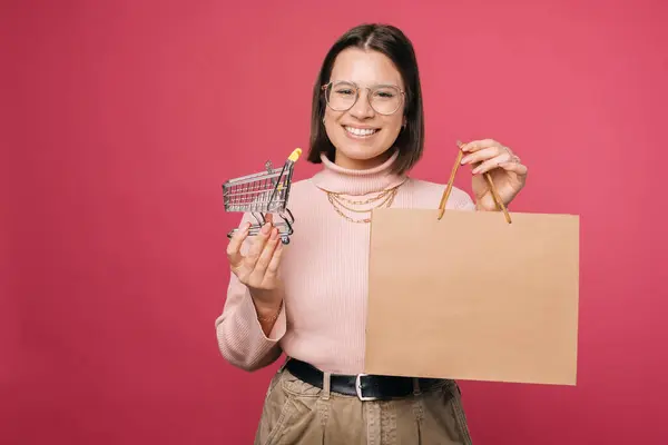 Молодая улыбающаяся женщина держит переработанную бумажную сумку для покупок и маленькую тележку. — стоковое фото