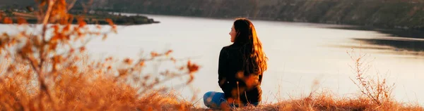 Młoda kobieta siedzi sama na polu i patrzy na rzekę. — Zdjęcie stockowe