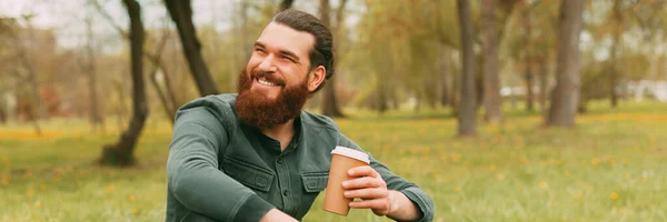 Портрет усміхненого бородатого чоловіка, який сидить у парку на траві і п'є чашку кави, щоб піти — стокове фото