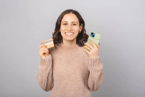 Atractiva jovencita sostiene un teléfono y una tarjeta de crédito sobre fondo gris. — Foto de Stock