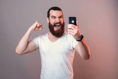 Güçlü sakallı adam telefon ekranına bakarken yüksek sesle bağırıyor. Gri arkaplanda portre.