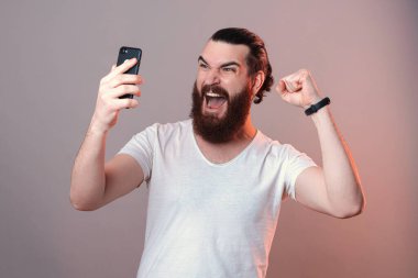 İçerik sakallı adam telefon ekranına bakarken çığlık atıyor ve kazanan jesti gri arka planda yapıyor..