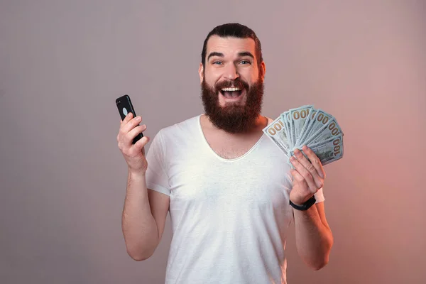 兴奋的大胡子男人手里拿着一堆钱和他的智能手机 在灰蒙蒙的背景上闪烁着红光 — 图库照片