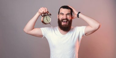 Genç sakallı adam kafasına dokunup çığlık atarken çalar saati tutuyor. Stüdyo görüntüsü gri arkaplan üzerinde kırmızı ışıkla.