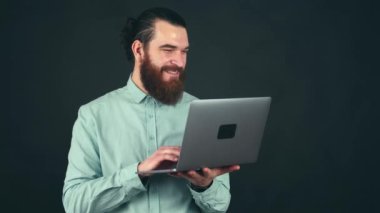 Laptop 'ta çalışan sakallı genç bir adamın karanlık arka planda çalışması ve gülümsemesi.