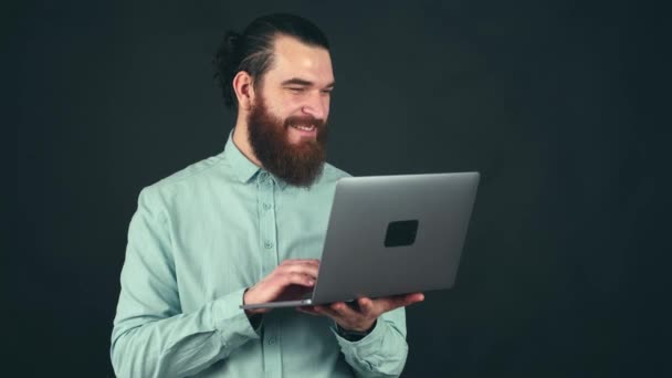 Nagranie młodego brodatego biurowca pracującego na laptopie na ciemnym tle i uśmiechającego się — Wideo stockowe