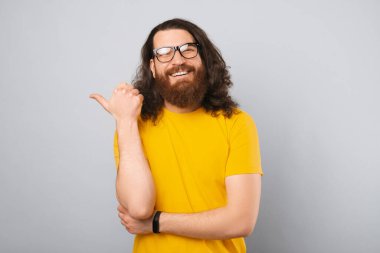 Uzun saçlı sarı tişört giyen heyecanlı sakallı adam baş parmağıyla işaret ediyor. Stüdyo çekimi gri arkaplan üzerinde.
