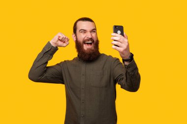 Kendinden geçmiş güçlü sakallı adam, sarı arka plandaki akıllı telefonuyla bir stüdyoda selfie çekiyor.. 