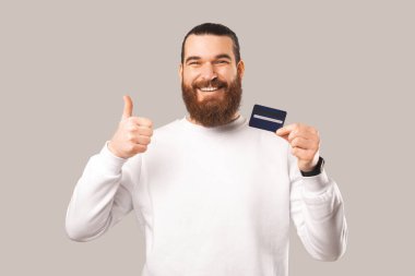 Ekstatik sakallı adam kredi kartı tutarken başparmağını kaldırıyor. Stüdyo çekimi beyaz arkaplan üzerinde.