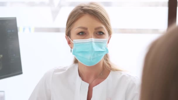 Nahaufnahme einer jungen Zahnärztin, die eine Maske trägt und mit einer Patientin in der Klinik spricht — Stockvideo
