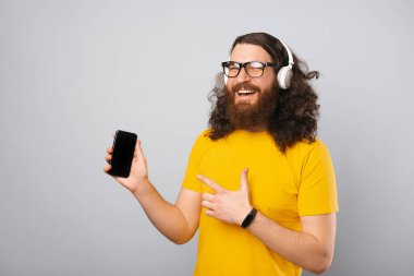 Sarı tişört giyen sakallı adam kulaklıklardan ve telefondan müzik dinlemekten zevk alıyor ve onu işaret ediyor. Stüdyo çekimi gri arkaplan üzerinde.