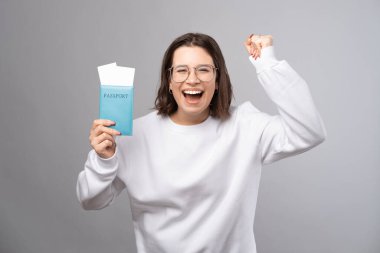 Kendinden geçmiş genç bayan, elinde pasaport ve gri arka planda dans ederken macerasından dolayı heyecanlı..