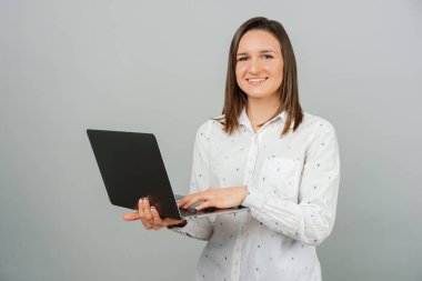 Neşeli kadın gri arka planda dururken elinde bir dizüstü bilgisayar tutuyor. Stüdyo çekimi.